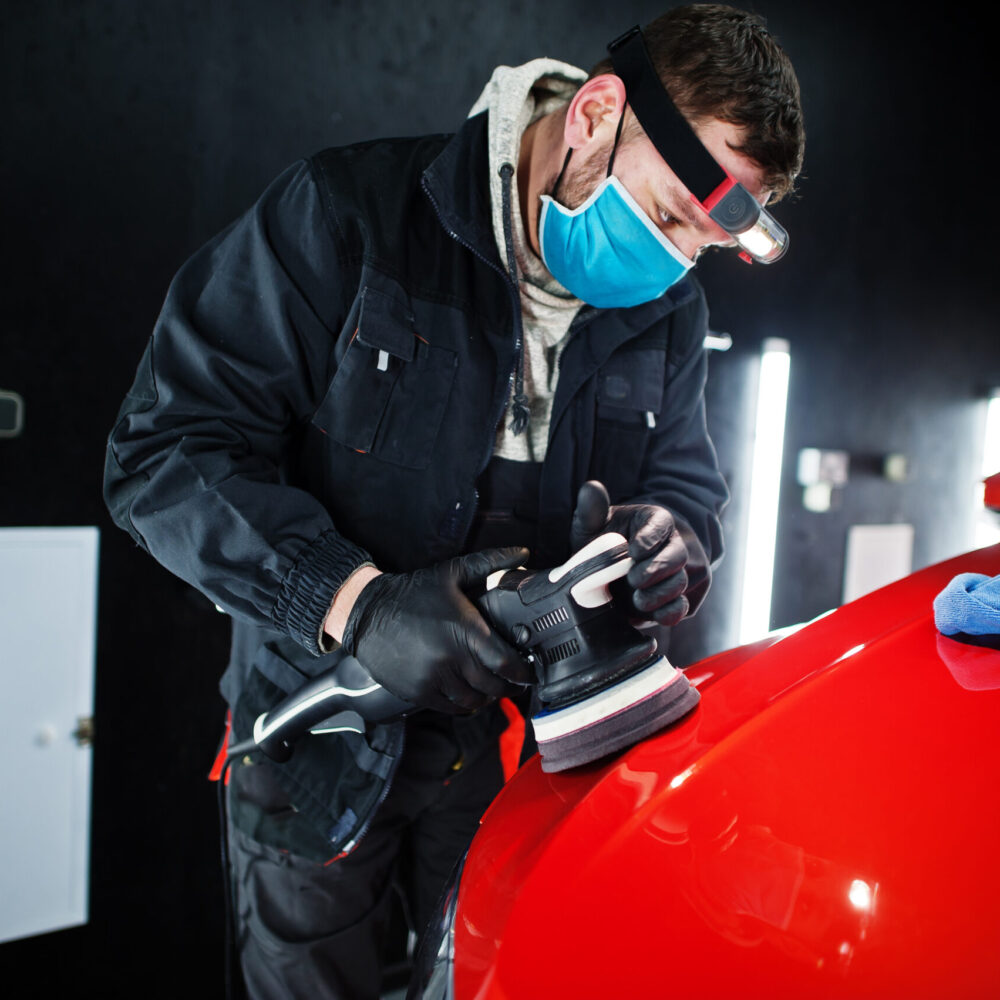 Car detailing concept. Man in face mask with orbital polisher in repair shop polishing orange suv car.