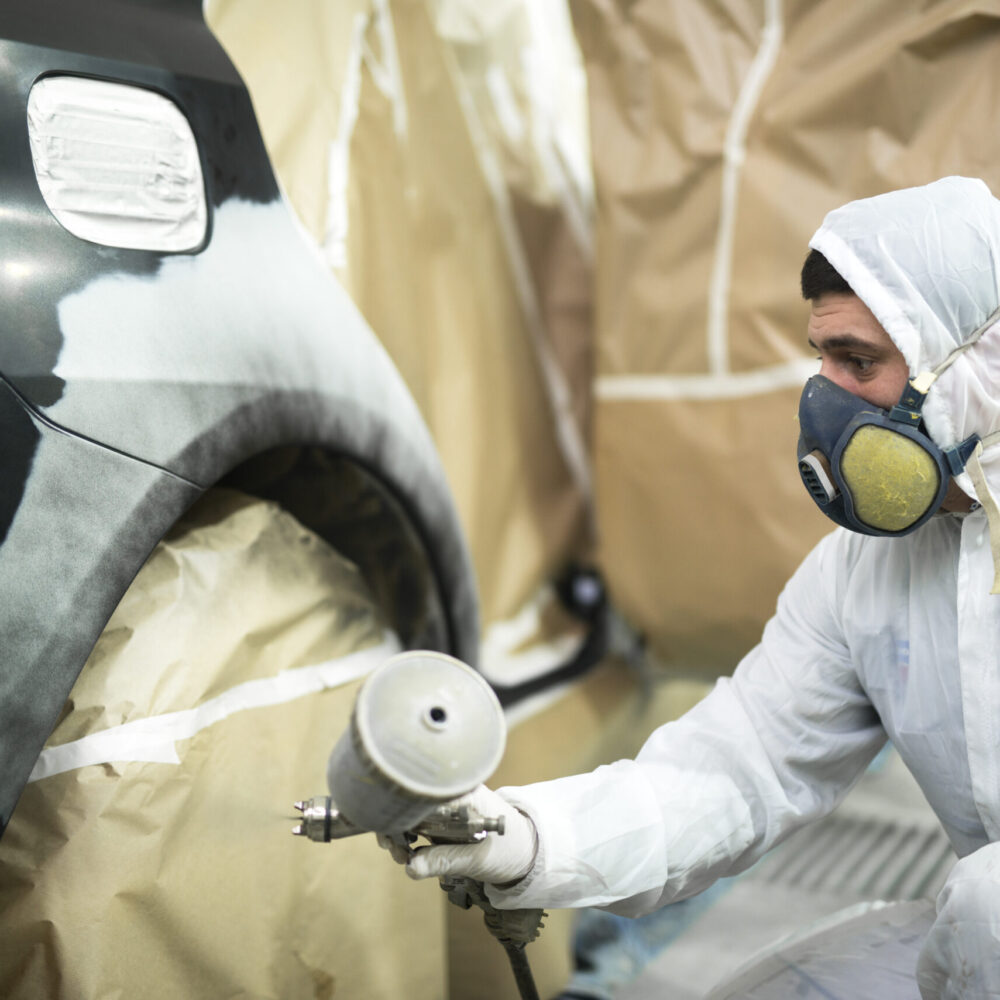 Man with protective clothes and mask painting automobile car bumper in repair shop.
